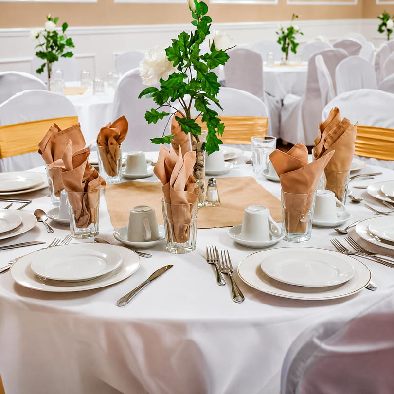 Elegantly set round tables with white tablecloths, gold sashes, and floral centerpieces in a banquet hall.