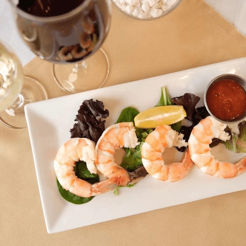 Shrimp cocktail with lemon, greens, and sauce on a white plate, with wine glasses nearby.