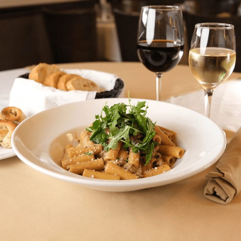 Pasta with arugula and parmesan, bread basket, and two glasses of wine on a table.