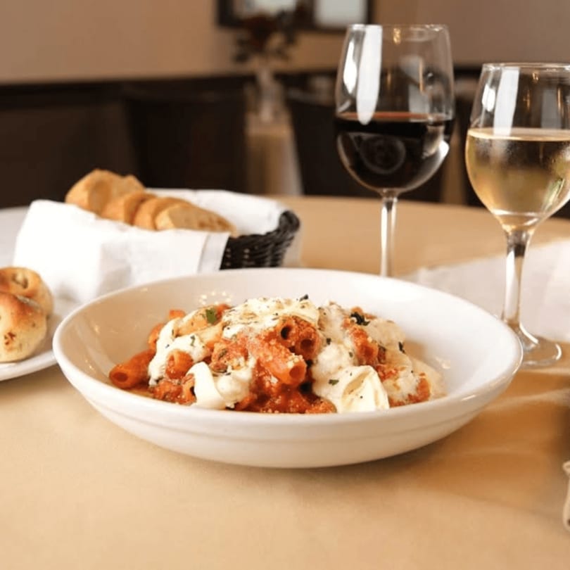 Pasta with red sauce and melted cheese, bread, and wine glasses on a restaurant table.