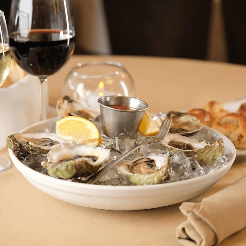 Fresh oysters on ice with lemon wedges and cocktail sauce, alongside wine glasses on a table.