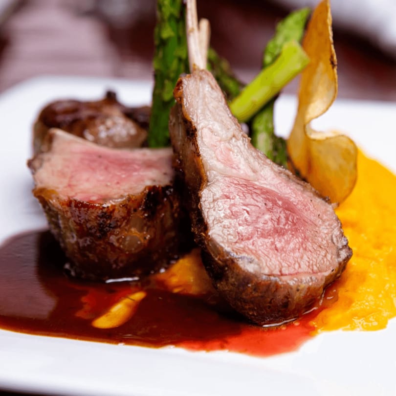 Two lamb chops with asparagus, potato crisp, and sauces on a white plate.