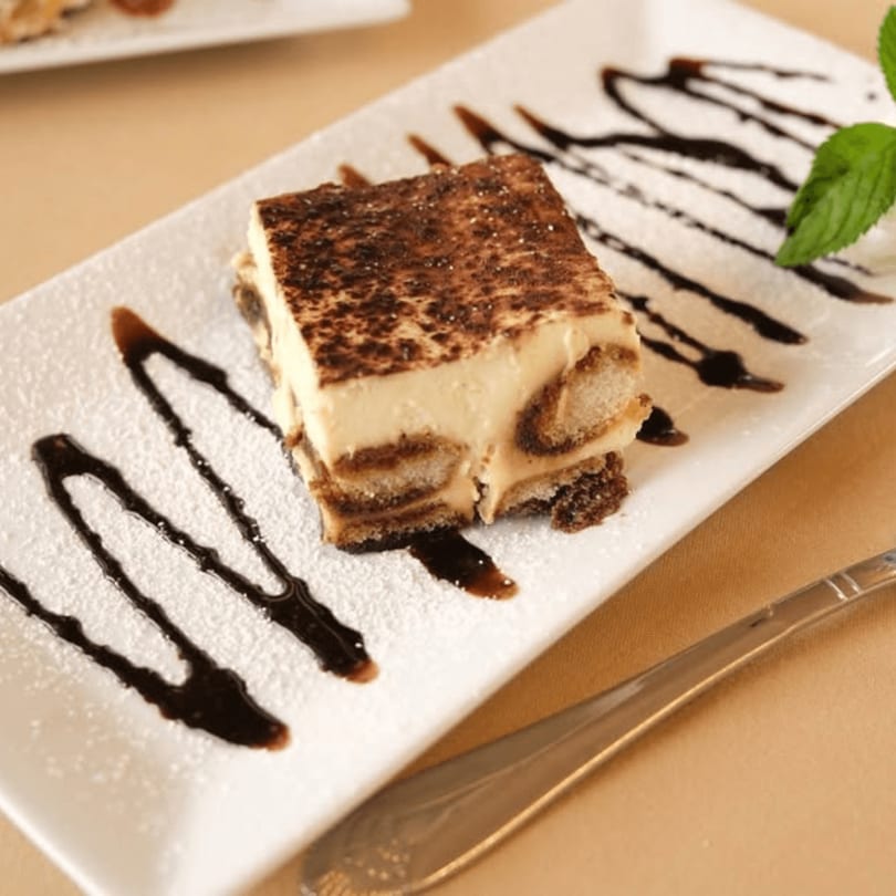 A slice of tiramisu dessert with chocolate drizzle and a mint leaf on a white rectangular plate.